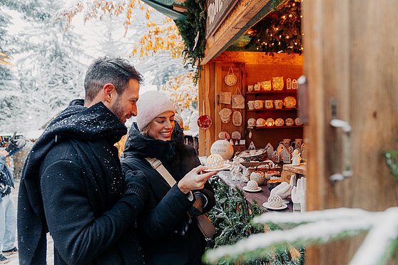 Tourismusverband Donau Oberösterreich | CM Visuals: Ein Paar betrachtet liebevoll handgefertigte Teelichthalter an einem Stand auf der Waldweihnacht Kopfing, verzaubert von ihrer Schönheit und der warmen Atmosphäre, die sie versprechen.