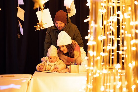 Tourismusverband Donau Oberösterreich/Studiosoco GmbH:
Ein Mädchen schreibt mit der Mutter einen Wunschbrief, während der Vater lächelnd dahinter steht.